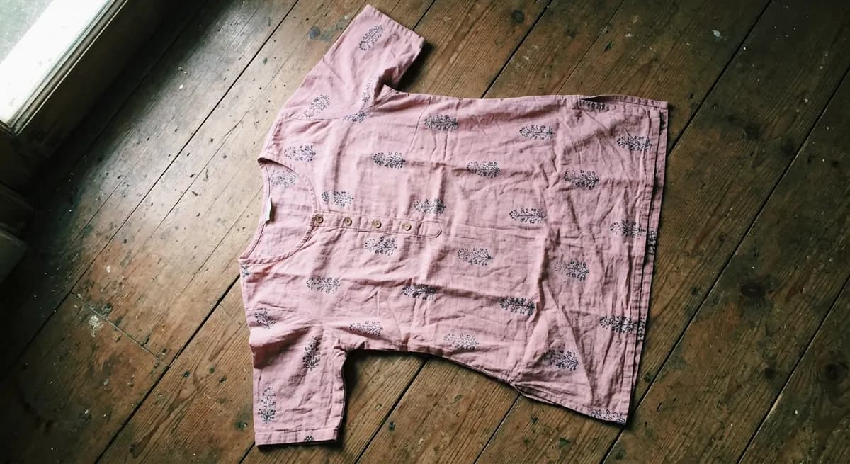 Amateur phone camera flat-lay of pale pink cotton kurta on wooden floor — typical seller product photo before AI photoshoot